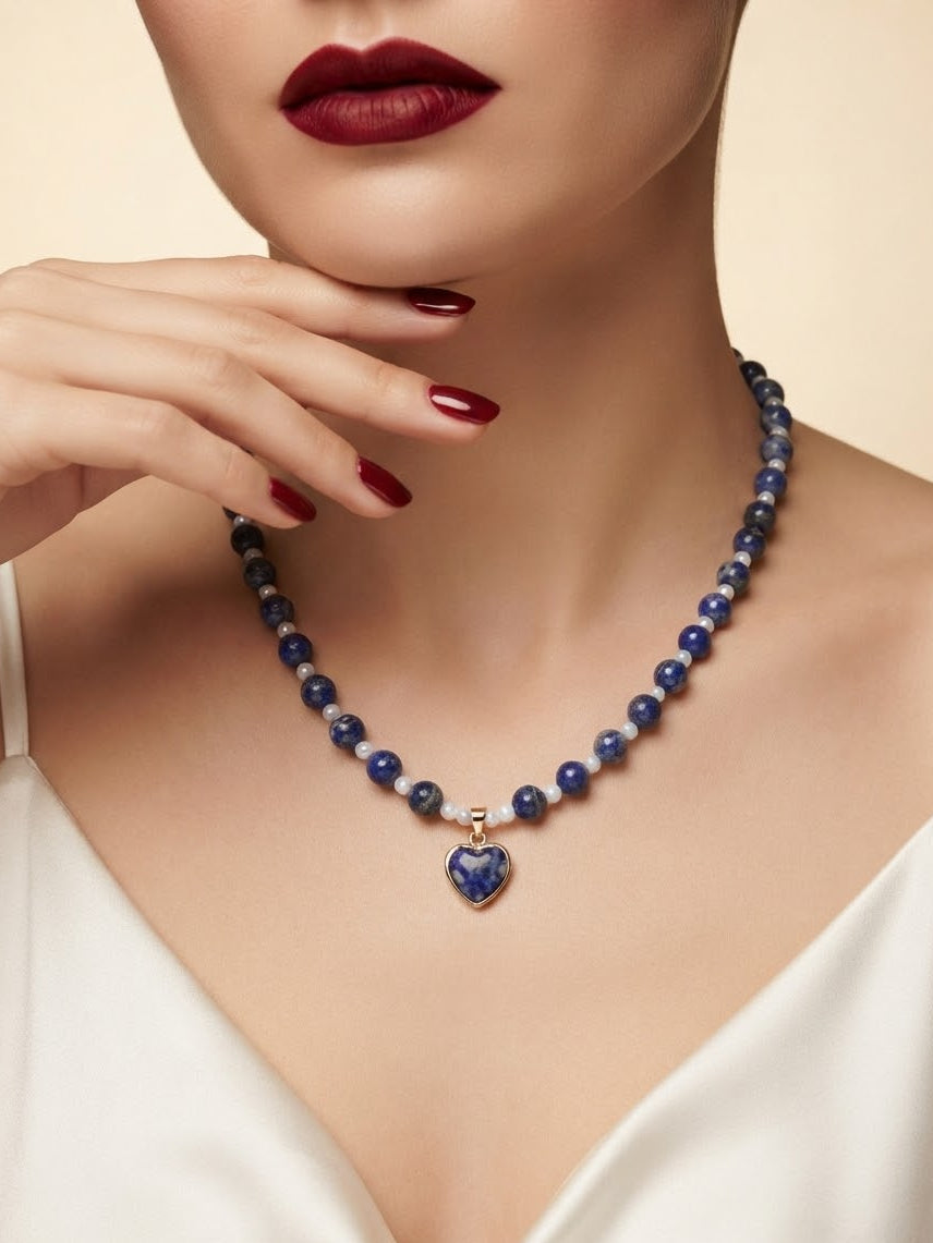 A close-up of a model wearing a beaded necklace with dark blue and white beads and a heart-shaped pendant, paired with deep red lipstick and white satin clothing.