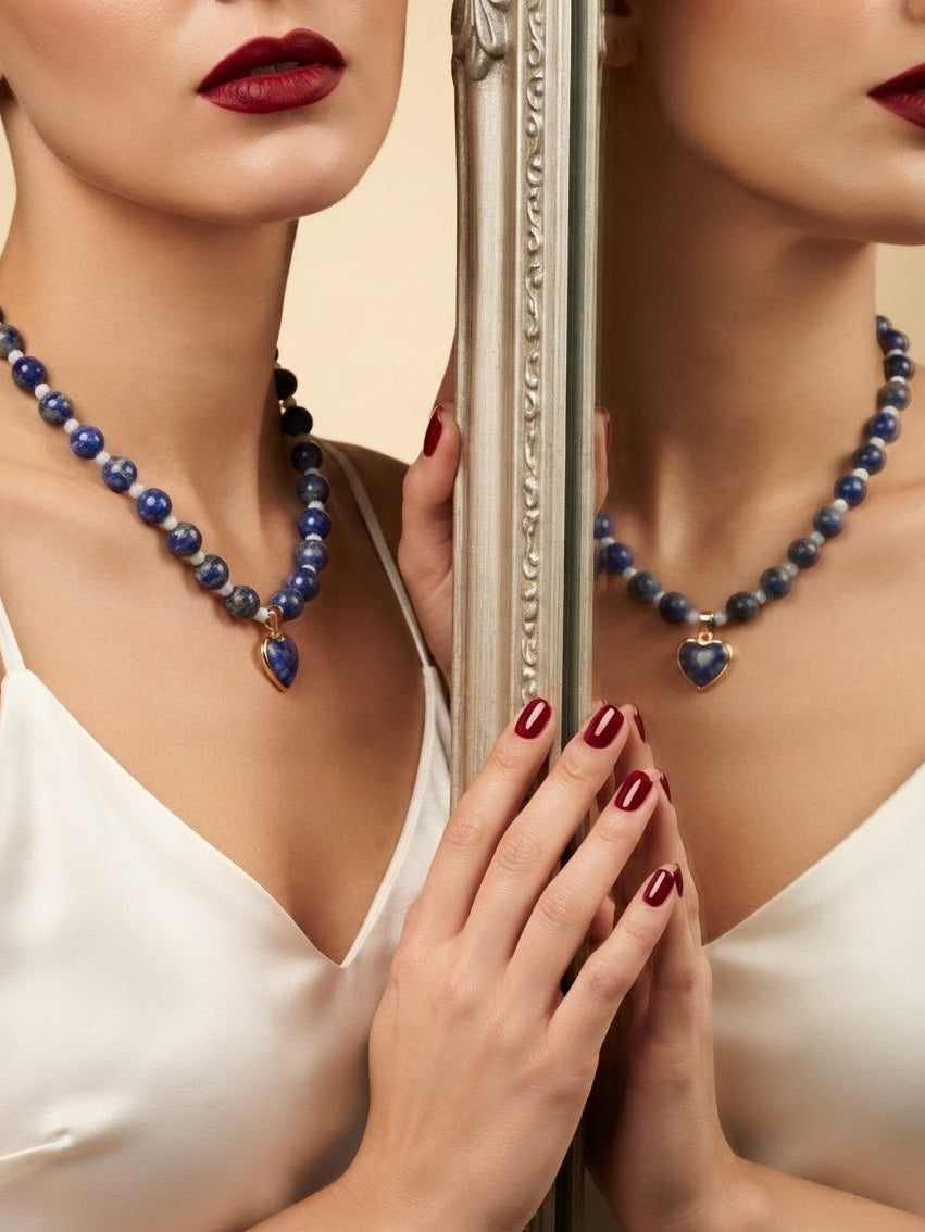 A close-up lifestyle shot of a model wearing a beaded necklace with blue and white beads and a heart-shaped pendant, posed in front of a mirror showing her reflection.