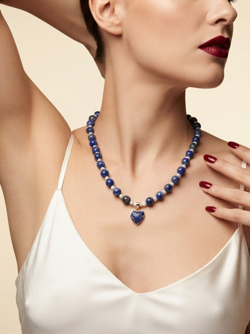 A close-up of a model wearing a beaded necklace made of deep blue lapis lazuli stones with a heart-shaped pendant, paired with a white satin top.