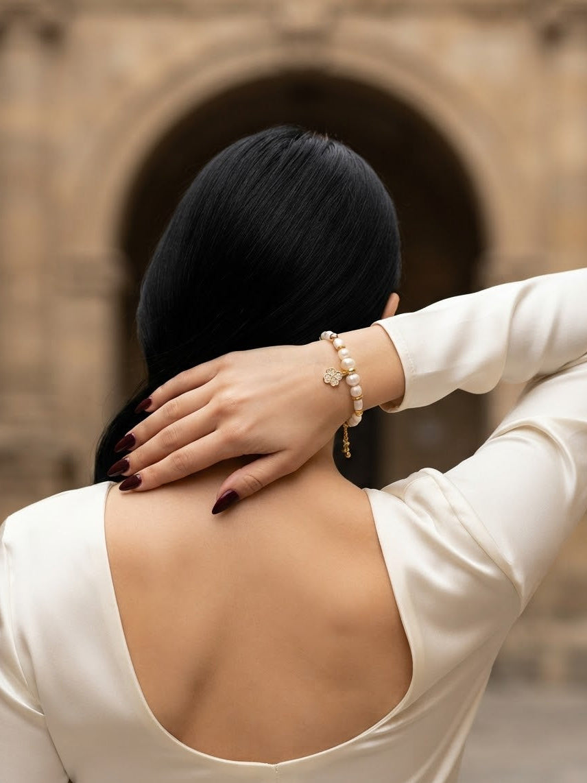A woman viewed from behind wearing a cream satin long-sleeve top, showcasing an open back. She is wearing a delicate pearl and gold beaded bracelet on her wrist.