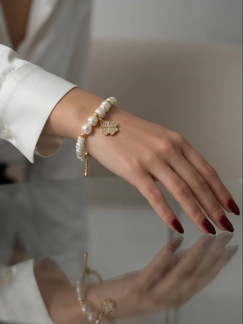 A close-up of a person's hand wearing a pearl bracelet with a gold clover charm and a small gold chain, paired with a white long-sleeve blouse.