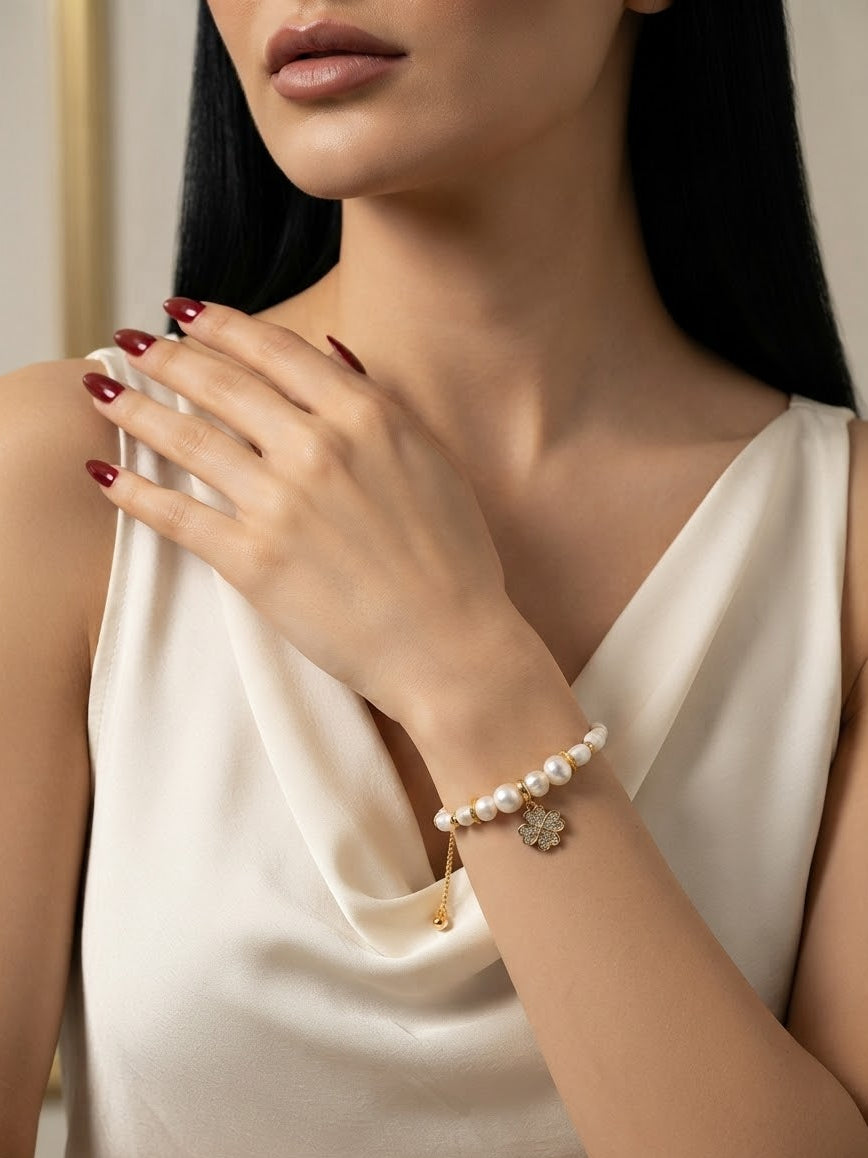 A close-up of a woman wearing a delicate pearl bracelet with gold charms, including a small flower charm, styled with a cream satin top.