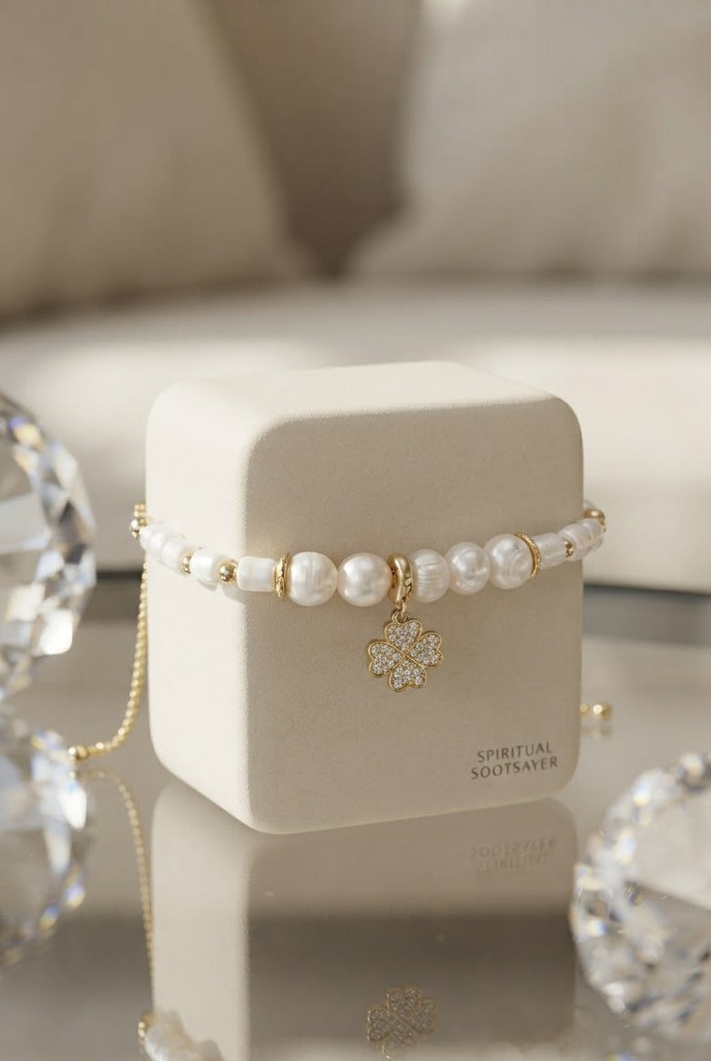 A beaded bracelet with white pearls and gold-toned beads, featuring a gold-toned crystal clover charm, displayed on a cream-colored jewelry box.