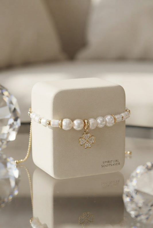 A beaded bracelet with white pearls and gold-toned beads, featuring a gold-toned crystal clover charm, displayed on a cream-colored jewelry box.