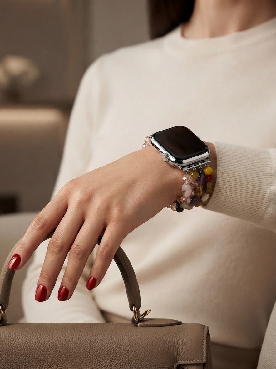 A close-up of a person's hand wearing a silver smartwatch with a multi-strand beaded bracelet watch band, paired with a cream sweater and holding a taupe leather handbag.