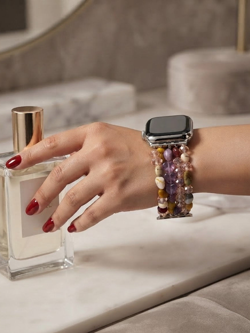 A close-up of a person's hand with red nail polish resting on a perfume bottle next to a smartwatch featuring a colorful beaded crystal band.