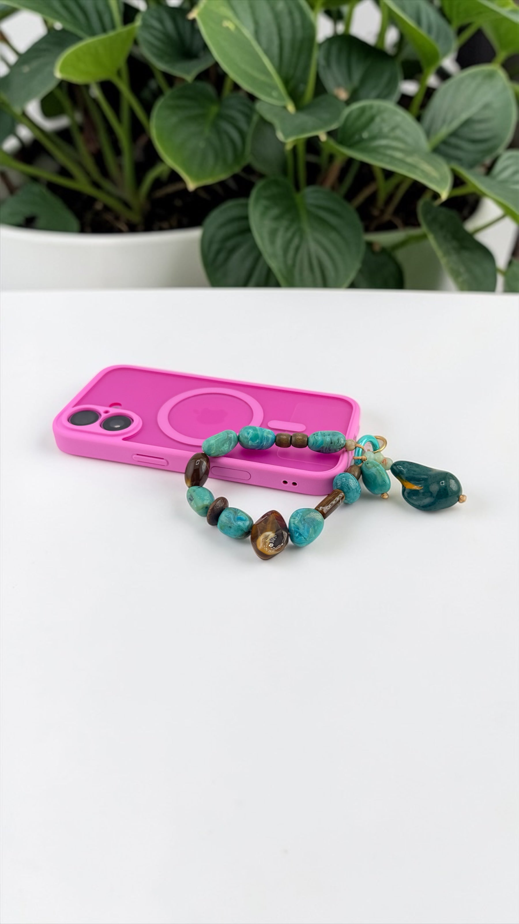 Pink phone case with green beads on a white surface, plant in the background