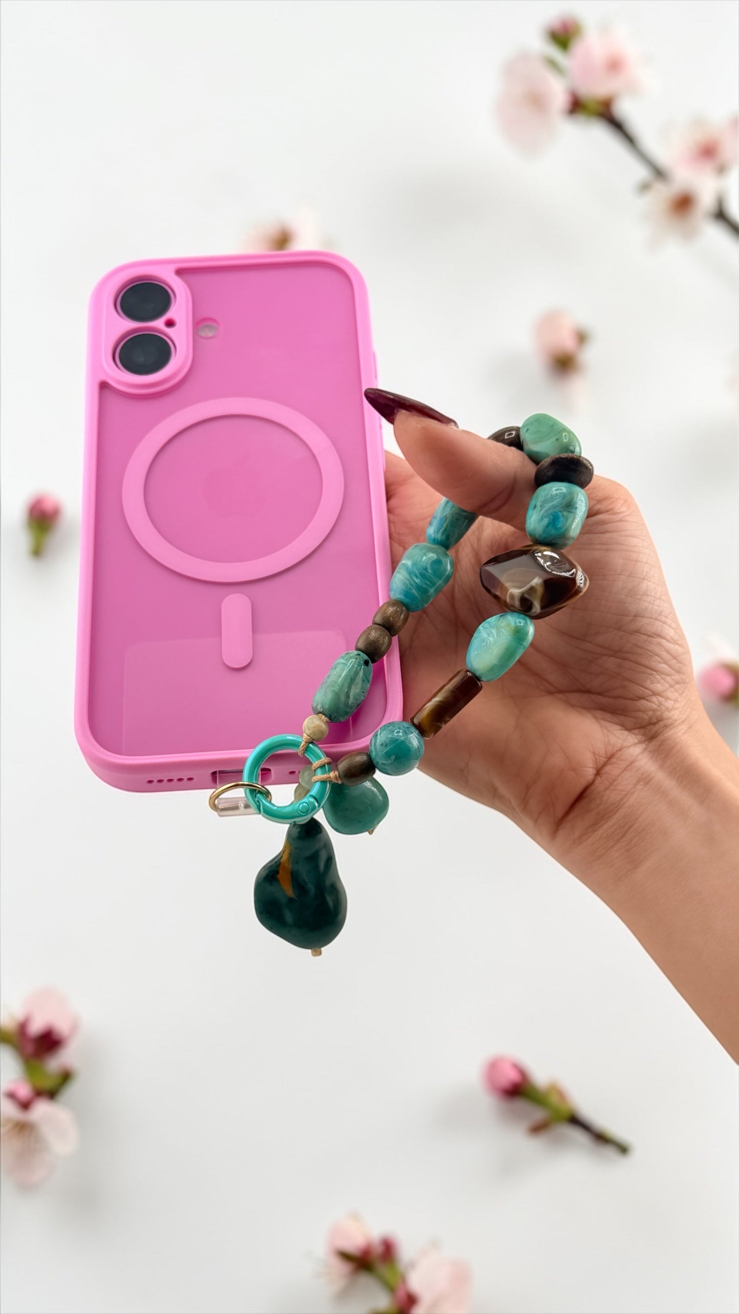 Hand holding a pink phone case with a turquoise charm against a light background with floral elements.