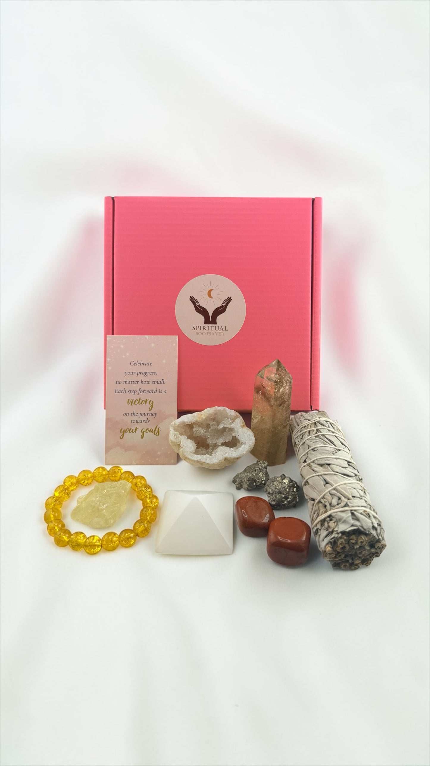 Pink box with a logo, surrounded by crystals, candles, and a bracelet on a white background