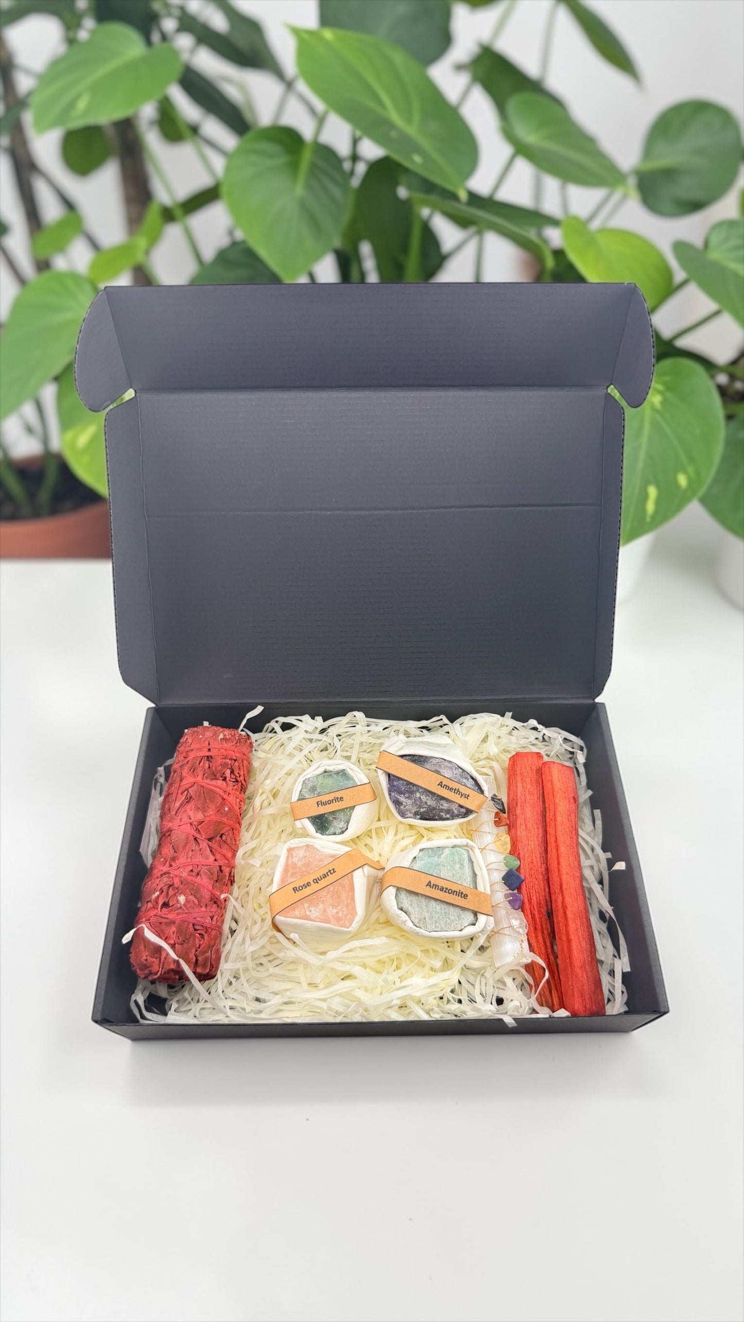 Decorative set with a mask, candles, and ribbons in an open black box against a green plant background.