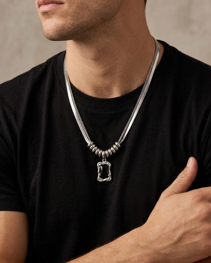 A close-up of a man wearing a silver chain necklace with a textured rectangular pendant over a black t-shirt, set against a neutral textured gray background.