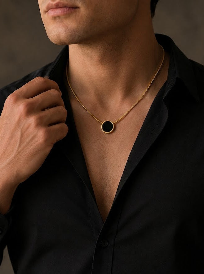 Man wearing a black shirt with a gold necklace featuring a black circle pendant against a neutral background