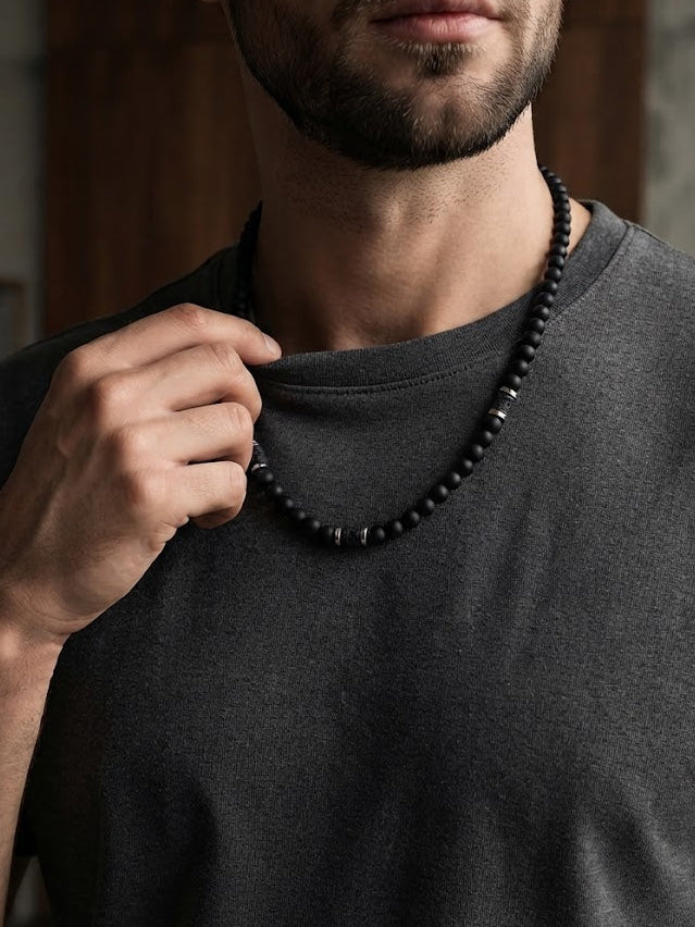 A close-up of a man wearing a dark charcoal gray t-shirt and a black beaded necklace, with his hand adjusting the collar of his shirt.