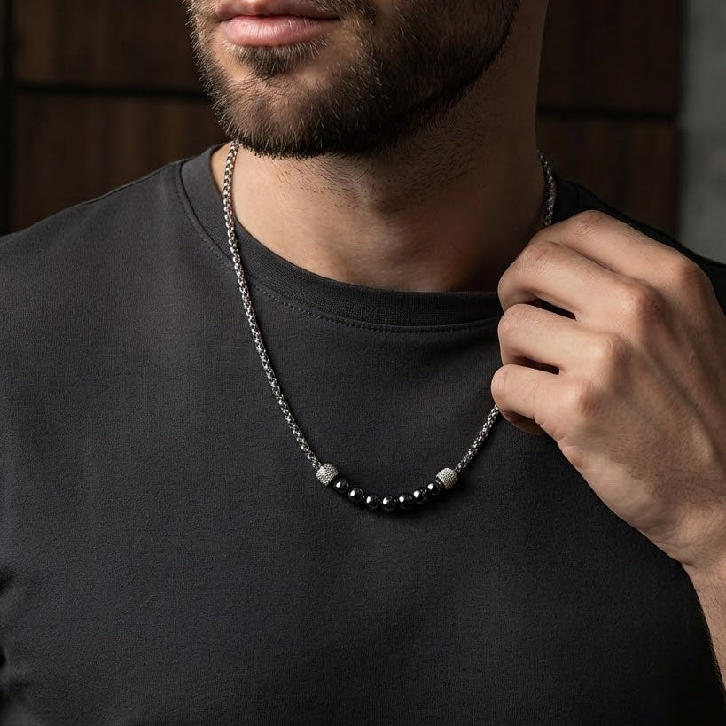 A close-up of a man wearing a silver-toned beaded necklace with a dark t-shirt, adjusting the chain with his hand.