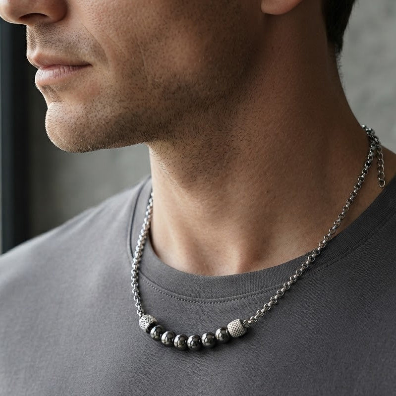 A close-up of a man wearing a silver-toned beaded necklace with a textured central bead, styled with a dark gray crewneck t-shirt.