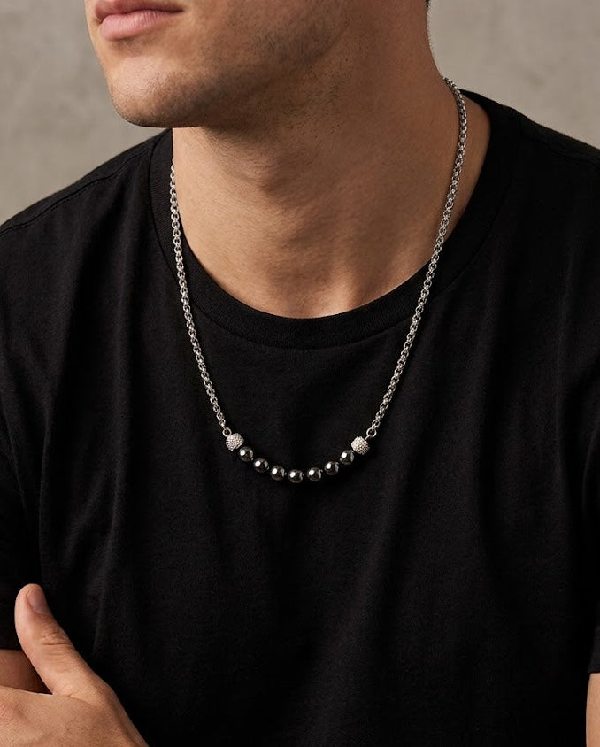 A close-up of a man wearing a silver chain necklace with a central cluster of dark beads and two lighter round beads, styled with a black t-shirt against a textured gray wall.