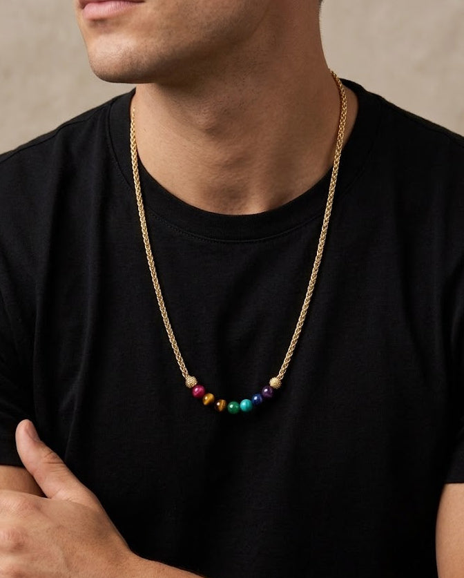 A man wearing a gold chain necklace with a small row of colorful gemstone beads at the center, paired with a black t-shirt against a neutral textured background.