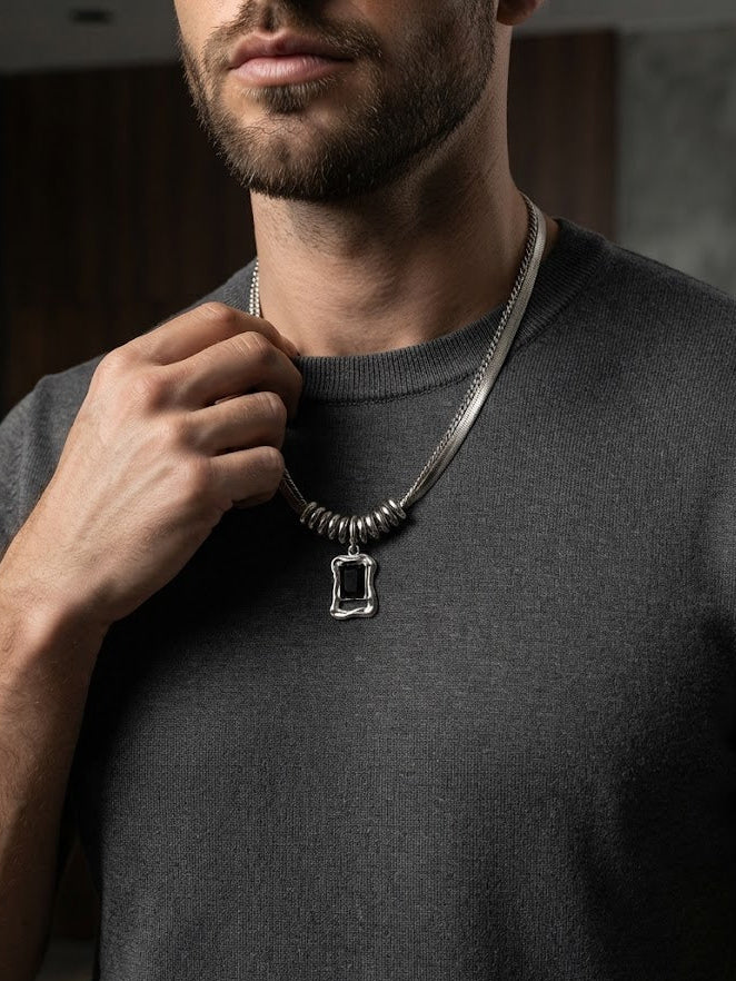 A close-up of a man wearing a silver-toned chain necklace with a rectangular bezel pendant, paired with a dark gray t-shirt.