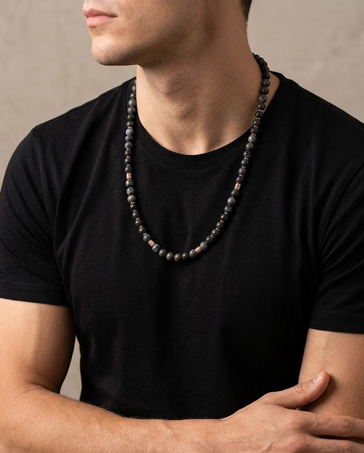 Man wearing a black t-shirt and a beaded necklace against a neutral background
