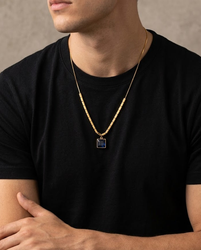 Man wearing a black t-shirt and gold necklace with a blue pendant against a neutral background