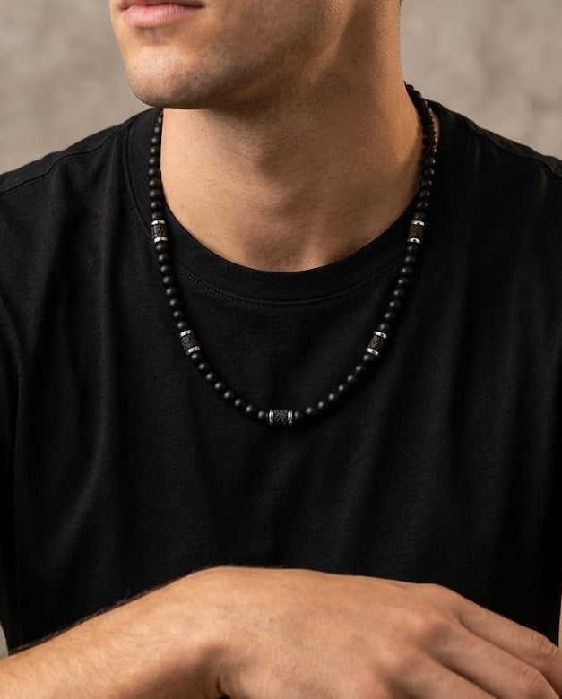 A close-up of a man wearing a black beaded necklace with silver-toned spacer beads and a black pendant, paired with a black t-shirt against a textured gray background.