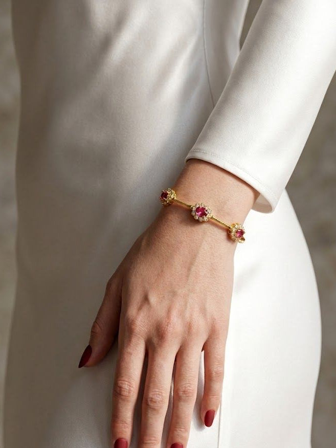 A gold bracelet with three pink gemstone accents worn on a model's wrist, shown against a white dress and a neutral background.