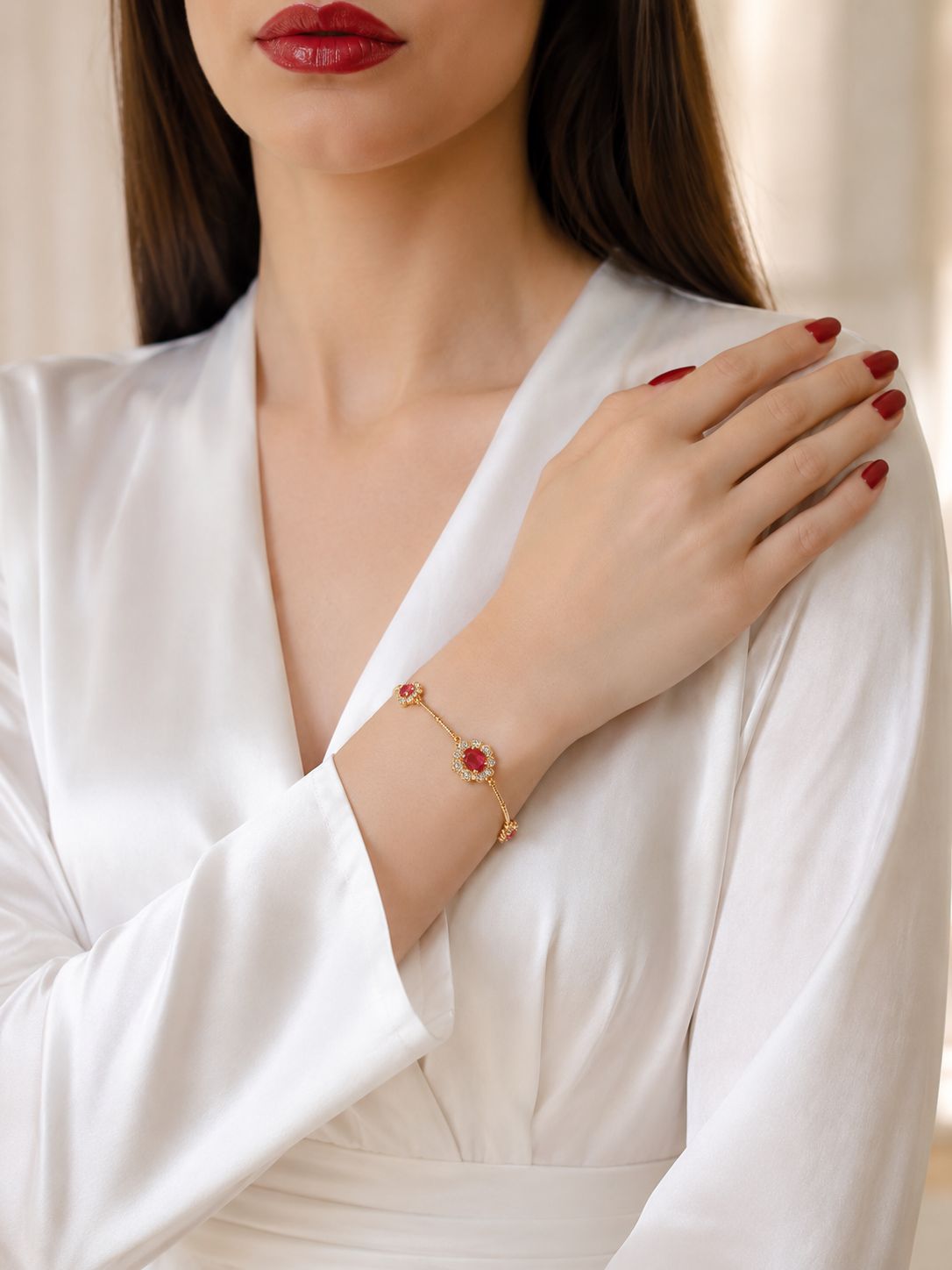 A close-up of a woman wearing a delicate gold bracelet with red oval gemstones, styled with a white satin robe and red nail polish.