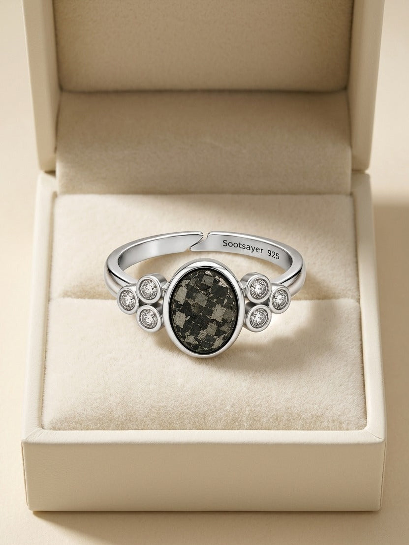 A silver-toned ring with a central dark marbled oval stone and small clear stones on either side, displayed inside an open cream jewelry box. The inside of the band is engraved with 'Scottyler 325'.