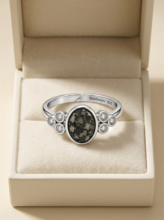 A silver-toned ring with a central dark marbled oval stone and small clear stones on either side, displayed inside an open cream jewelry box. The inside of the band is engraved with 'Scottyler 325'.