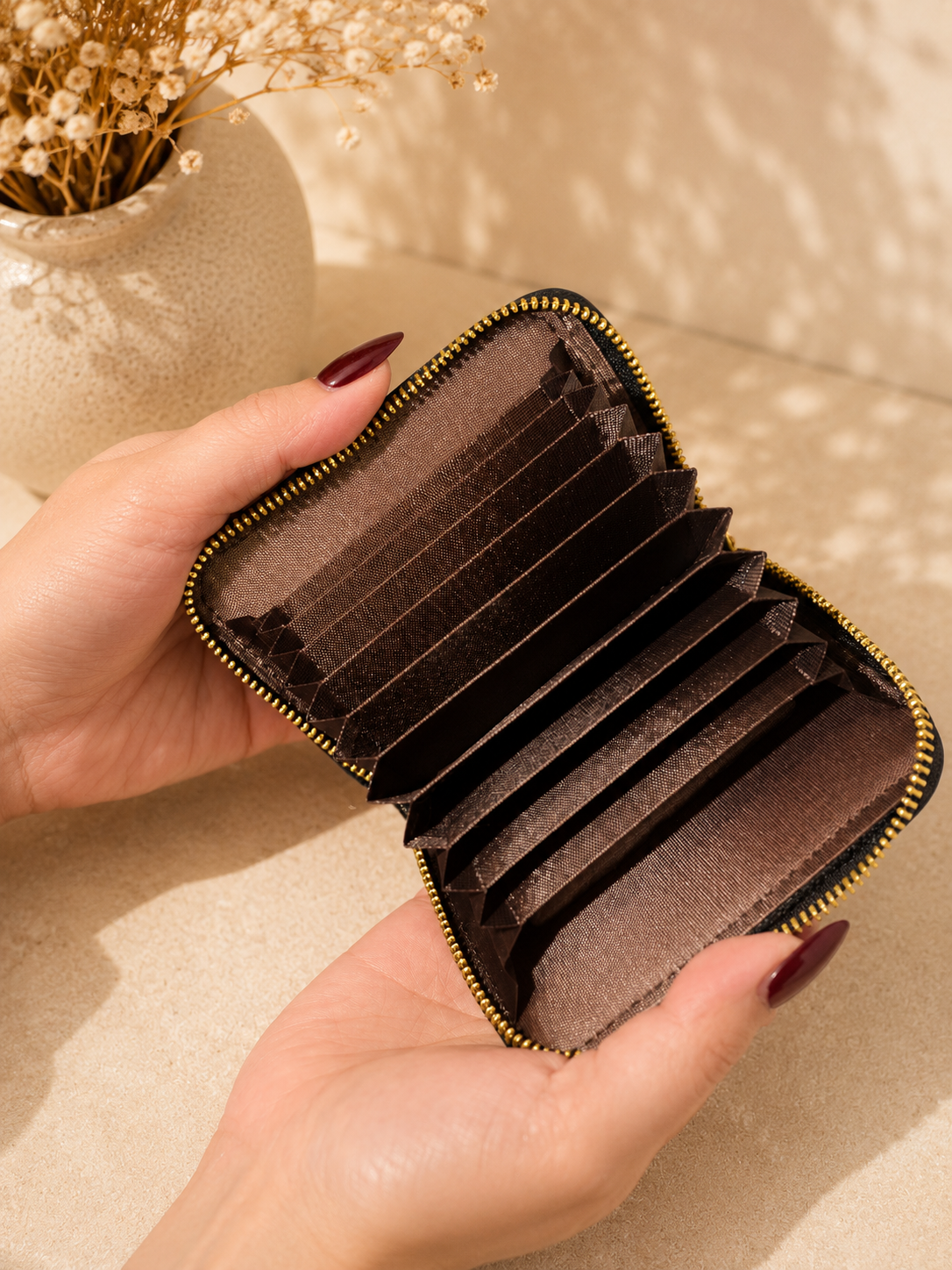 Brown cardholder with gold zipper held by a hand against a beige background