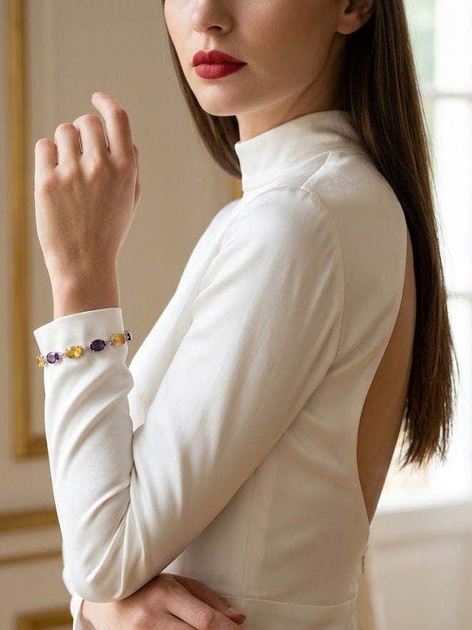 A woman wearing a white mock-neck long-sleeve top with an open back, showcasing a delicate bracelet on her wrist. The bracelet features a row of small oval beads in purple, yellow, and gold.