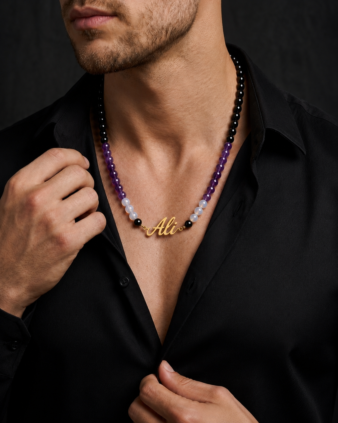Man wearing a black shirt with a beaded necklace and gold nameplate, against a dark background.